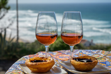 Portugal's traditional food and drink, glasses of porto wine or muscatel de setubal and dessert Pastel de nata egg custard tart pastry served with view on blue Atlantic ocean in Lisbon area, Portugal