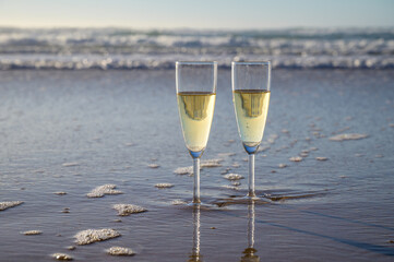 Glasses of cava or champagne sparkling wine on white sandy ocean beach with water waves on sunset in sunlights