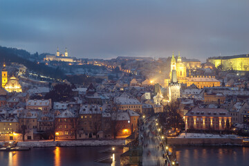 Blick über die Karlsbrücke über der Moldau in Prag auf die Burg und den Petrin am Abend, im Winter, im Schnee, mit erleuchteten Häusern und Menschen