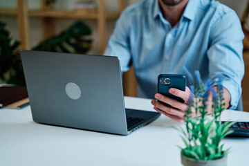 Young male freelancer in casual clothes sitting at table with laptop and browsing mobile phone while working on remote project at home