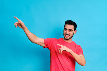 Naklejka premium Positive young bearded male in red shirt and white shirt pointing away while standing against blue background and looking at camera