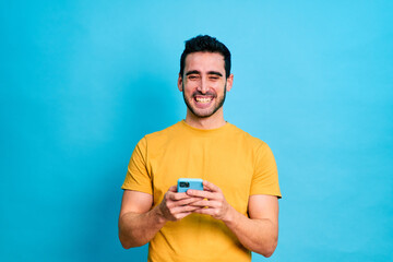 Young bearded male in casual clothes browsing mobile phone while standing against blue background