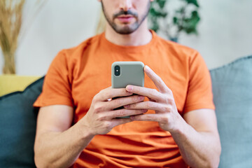 From above of crop anonymous male in red shirt browsing mobile phone while sitting on sofa in living room at home