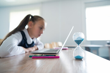 Caucasian girl sits at a desk at school and studies at a laptop. Hourglass on the table.