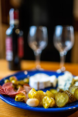 A plate filled with different kinds of cheese, olives, peppers, sausage, garlic and cucumber and a bottle of red wine with two glasses.