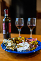 A plate filled with different kinds of cheese, olives, peppers, sausage, garlic and cucumber and a bottle of red wine with two glasses.