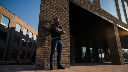 Caucasian bearded man in a bomber jacket leaned against a brick wall. 