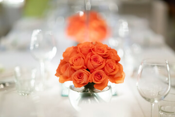 a large bouquet of red roses Looks outstanding in a glass vase in the center of the dining table. For guests waiting to sit and eat There were wine glasses and plates around.