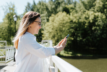 Teeth smiling young woman using smartphone outdoors. Side view positive caucasian woman wearing glasses browsing messages or internet on phone while standing on bridge in park