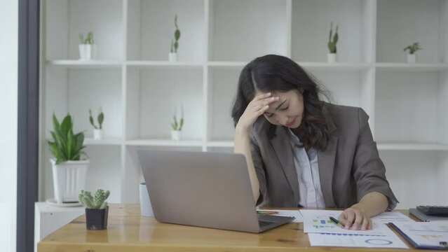 Asian businesswoman feeling stressed by working with laptop and financial accounting graph document Young woman tired at work in the office holding her head in her hands feeling headache