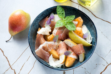Bowl with cantaloupe and pear slices, prosciutto, torn mozzarella and fresh basil, horizontal shot on a light-beige stone background