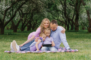 Happy beautiful family is sitting on a blanket in the park in the summer and looking at the blanket, talking via video link