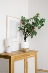 An artificial flower in a pot on a wooden chest of drawers in the living room, decorative items and a frame on the wall in a Scandinavian minimalist style