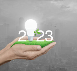 2023 white text and led light bulb with fresh leaves on green grass in hands over pollution of city tower and skyscraper, Happy new year 2023 green ecology and saving energy concept