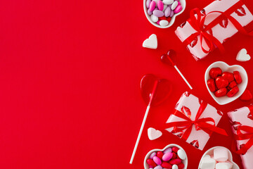 Love or Valentines concept. Top view composition made of gift boxes and heart shaped saucers with confectionery chocolate candies and lollipops on red background. Flat lay with copy space.
