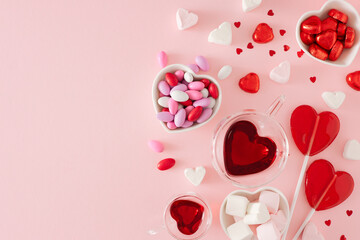 Valentines Day mood concept. Flat lay photo of heart shaped glass cup with drinking, chocolate candies and lollipops on pastel pink background with copy space. Sweet Valentines card idea.