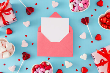 Love Valentines concept. Flat lay composition of gift boxes, heart shaped saucers with sweets candies and lollipops and marshmallow on blue background with paper envelope in the middle.