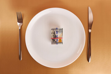A white plate with a roll of dollars, a fork and a knife on a golden background.