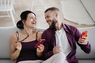 Cheerful hispanic woman sits on couch with husband enjoying vacations holding phone toothy smiles. Beard Caucasian young man in violet shirt looks at girlfriend tenderly. Holidays. Couple at cozy home
