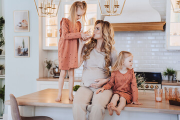 Pregnant caucasian mom sits on kitchen table with little daughters dressed in beige casual clothes. Daughter staying on the table holds a glass, helps her mom drink a milk. Maternity and pregnancy.