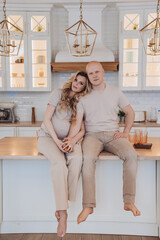 Vertical shoot of Swedish bald man in casual sitting with pregnant attractive wife on kitchen table, looks at camera. Happy female housewife leans on husbands shoulder at home. Parenthood. Couple home