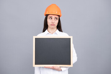 Young funny sad woman construction manager. Woman builder isolated portrait with protect helmet and board. Gray background, empty space, copy space.