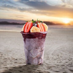 A cup of acai topped with strawberry and banana on a beach at sunset