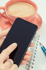 Hand of woman with mobile phone, coffee with milk and notepad for writing notes. White background