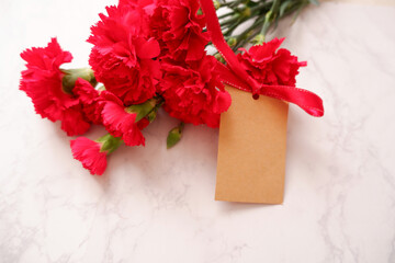 Red Carnation decoration with ribbon and blank message tag on white marble background. Carnation flower composition for Mother's day, Women's day and wedding celebration design. 