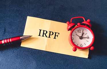 A sigla IRPF para Imposto sobre a Renda das Pessoas Físicas escrita em um pedaço de papel com um relógio na composição. Imposto de Renda no Brasil. - obrazy, fototapety, plakaty