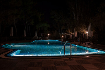 Folded parasols and empty deck chairs arranged by illuminated swimming pool with trees in the background at night in tourist resort