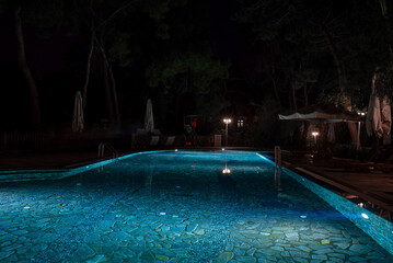 Parasols and empty deck chairs by illuminated swimming pool with trees in the background at night in tourist resort