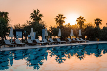 Empty deck chairs and folded parasols arranged around swimming pool with silhouette trees and clear sky in the background at tourist resort during sunset