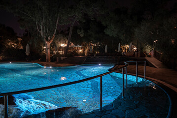 Metallic railing by illuminated blue swimming pool with trees growing in the background at night in tourist resort
