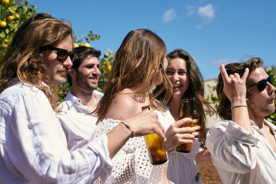 Group Of Happy Crazy Friends Having Fun Together Outdoors And Dancing. Women And Man Wearing White Clothes Chilling, Drinking On Party, Gesturing Horns By Fingers.