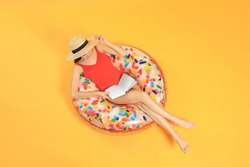 Happy young woman with beautiful suntan reading book on inflatable ring against orange background,...