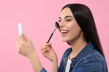 Beautiful woman with cosmetic pocket mirror applying makeup on pink background
