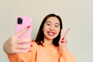 Cheerful Asian female in casual clothes smiling and showing sign while taking self portrait on...