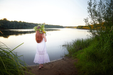 Fototapeta premium Portrait of beautiful slavic girl with long hair with flower crown near water of river or lake on nature in sunny evening. National selebration of summer holiday of Ivan Kupala