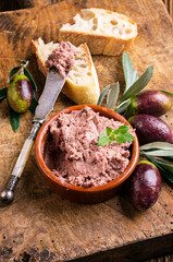 Traditional French goose pate as spread in a bowl with baguette and olives served as close-up on a rustic wooden board