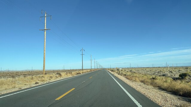 Wide View Desert Highway Leading To Distant Mountains, Yellow Markings On Asphalt, Telephone Poles On The Left And Blue Sky Overhead. California, Day Time