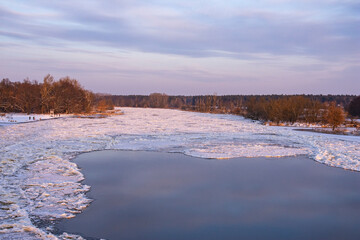 Ice-bound river and floating floe on a sunny frosty winter day. The shore is covered with trees and bushes. Winter.