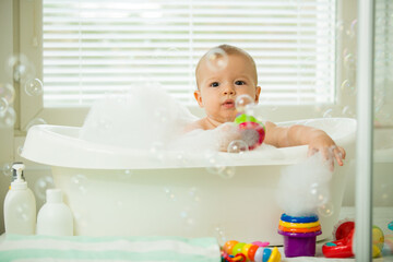Cute little baby sitting in white bathtub with foam and soap bubbles. Taking bath and playing with...
