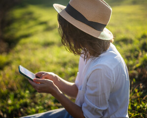 Fototapeta premium Happy pretty young cheerful woman in white shirt and sunglasses using smartphone while sitting on green grass on a sunny day
