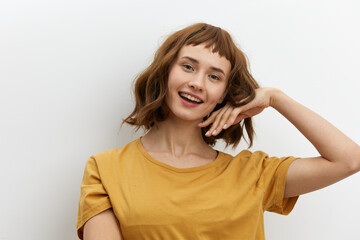 a beautiful, funny, attractive woman in a yellow cotton T-shirt stands on a light background and smiles broadly holding her hand near her beautiful well-groomed hair