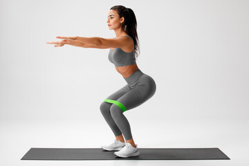 Athletic girl doing squats exercise for glute with resistance band on gray background. Fitness...