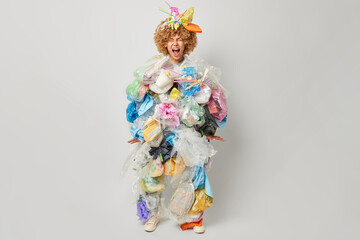 Studio shot of curly woman shouts loudly keeps mouth opened collects plastic rubbish tries to save planet from contamination isolated over grey background. Say no to plastic. Save world from rubbish