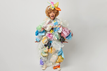 Full length shot of curly haired woman yells loudly made costume of plastic garbage takes care of environment isolated over grey background. Emotional female volunteer bothered by pollution.