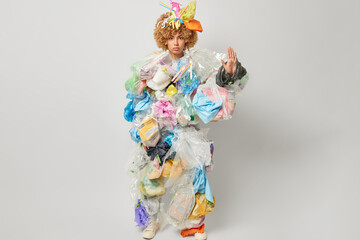 Photo of serious curly female volunteer makes stop gesture collects plastic garbage takes care of our planet asks to clean environment isolated over grey background. Global environment problem