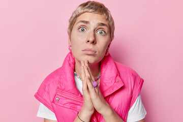 Sad desperate adult woman with short hair keeps palms pressed together begs you and asks for help makes wish apologizes for something dressed in fashionable clothes isolated over pink background - obrazy, fototapety, plakaty